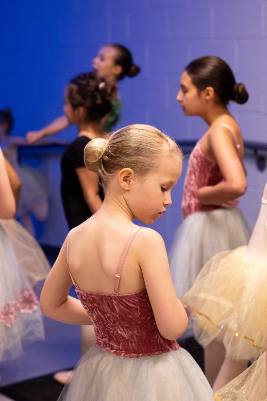 Young ballerina in pink and blue costume waits to backstage with other dancers before performing.